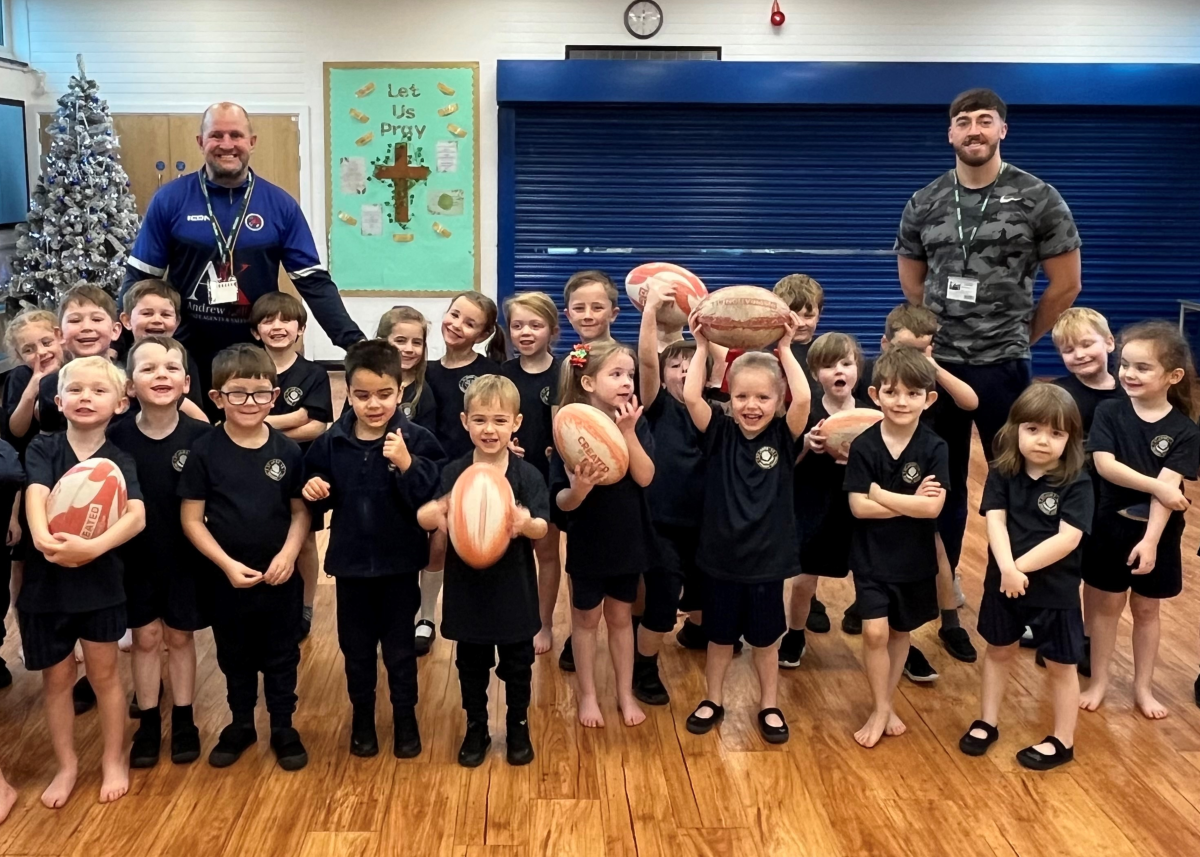 two male rugby coaches stood with large group of primary school aged children in PE kits in school hall