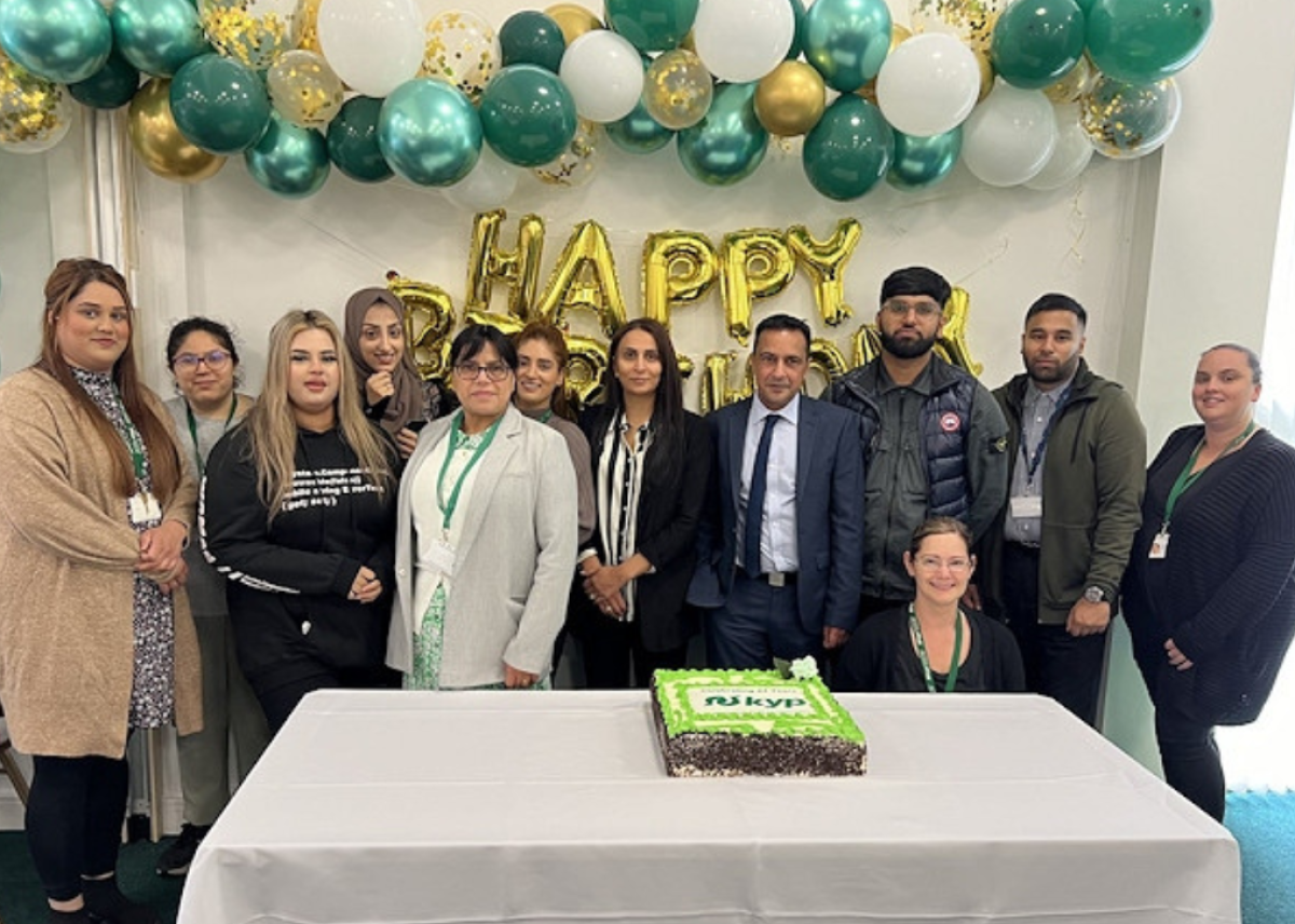 group of employees from KYP smiling in front of green, white and gold happy birthday balloons