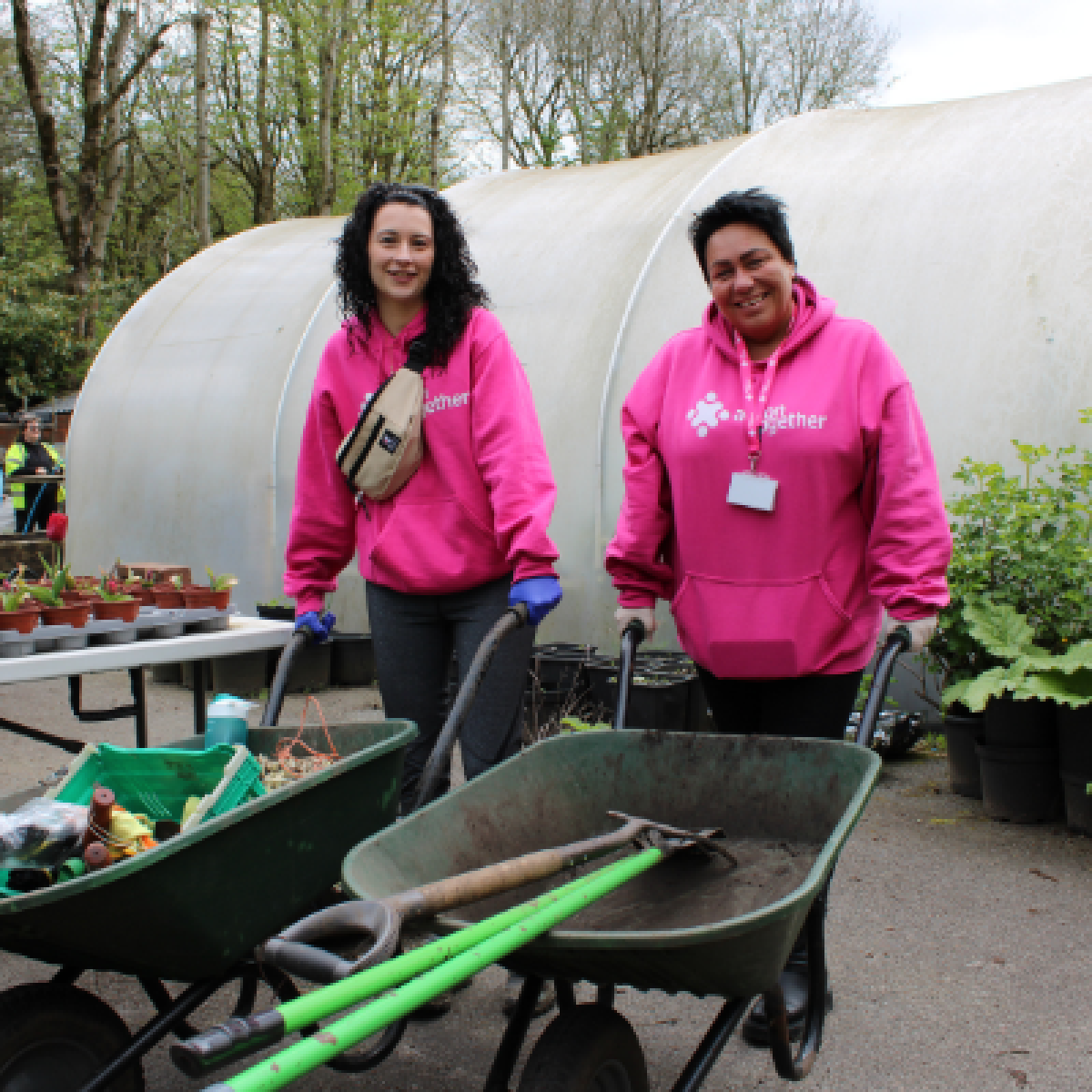 two women in bright pink hoodies pushing wheelbarrows in front of a large garden polytunnel