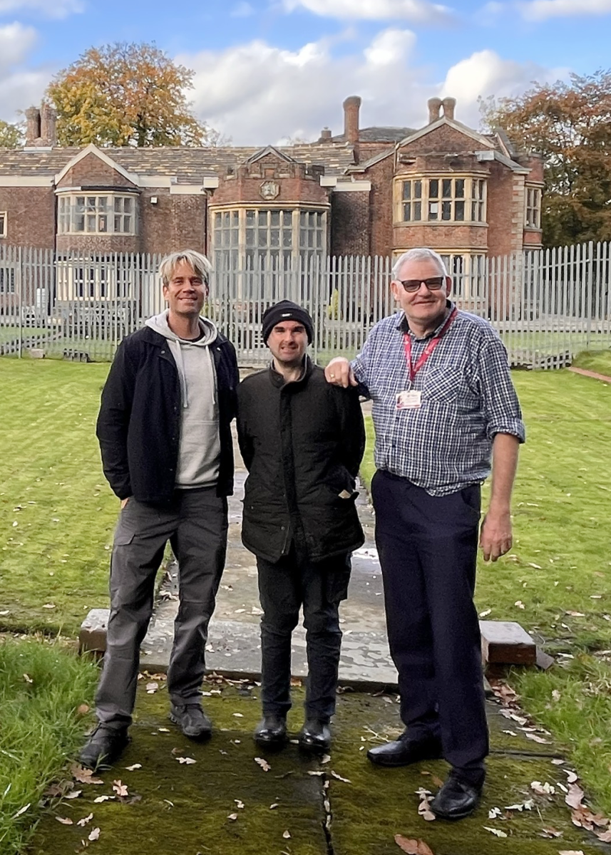 three men smiling at camera in front of old hall building-Phil G and volunteer Jon